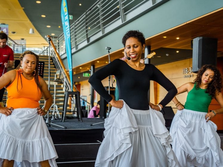 Image of Three women in colourful tops and white skirts perform a dance indoors on a stage, with a musician and microphones in the background. They have their hands on their hips and are smiling, mid-performance.