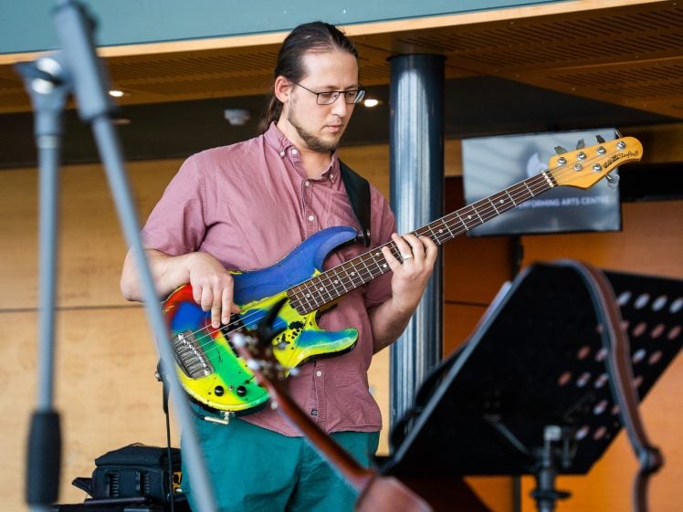Image of A man with glasses and a pink shirt plays a colourful electric bass guitar. He is focussed on his instrument, performing indoors, with a music stand and microphone visible in the foreground.