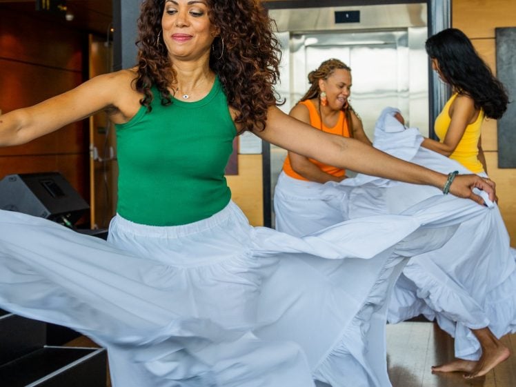 Image of Three women dance indoors, wearing bright sleeveless tops and long, flowing white skirts. The woman at the front spins, smiling, whilst the others dance joyfully in the background on a wooden floor.