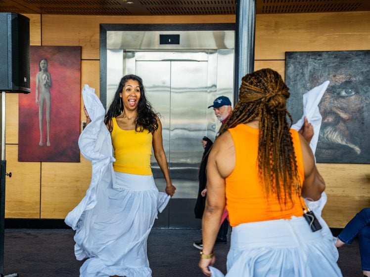 Image of Two women in yellow tops and white skirts dance energetically indoors. One faces the camera, smiling, whilst the other faces away. People and artwork, including portraits, are visible in the background.