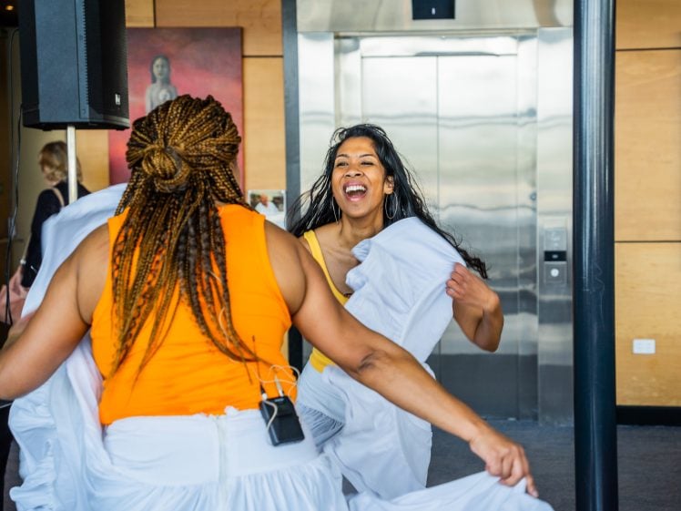 Image of Two women in matching orange tops and white skirts dance joyfully indoors, smiling brightly. One woman has long dark hair, while the other has plaits. A lift and wall art are visible in the background.