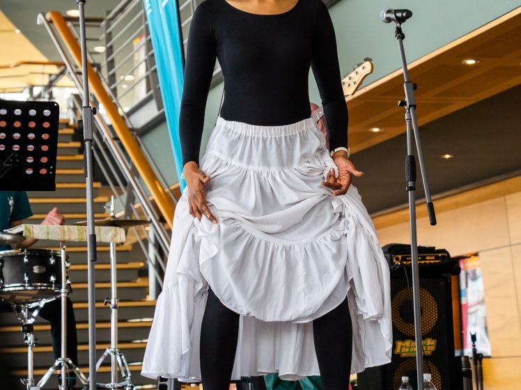 Image of A barefoot performer wearing a black top, white layered skirt, and headset microphone stands on stage in front of a staircase, with musical instruments and a blue banner in the background.
