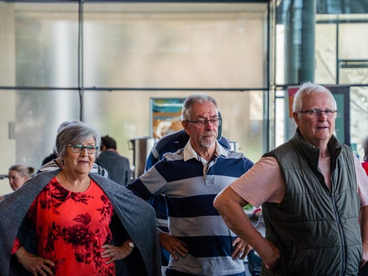 Image of Three elderly people stand indoors with hands on hips, appearing engaged and attentive. Two men and one woman are in the foreground, with other people and large windows in the background.
