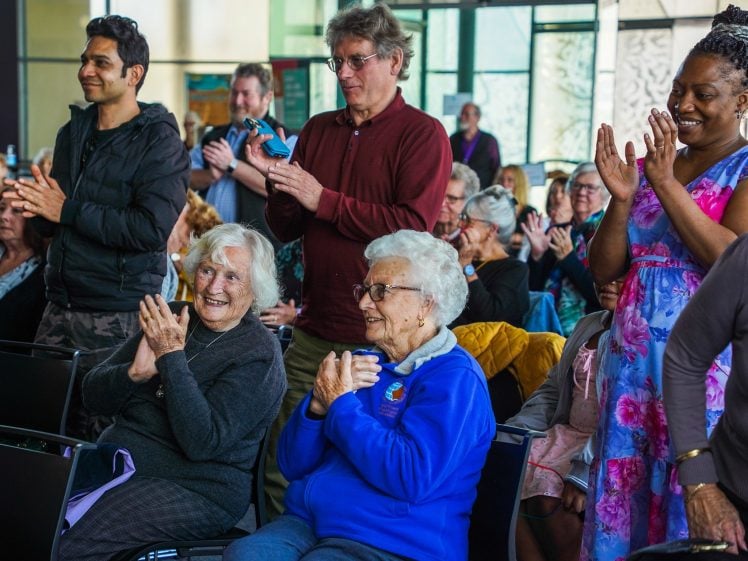 Image of A diverse group of people, including pensioners, smile and applaud whilst standing and sitting at an indoor event with natural light streaming in from large windows.
