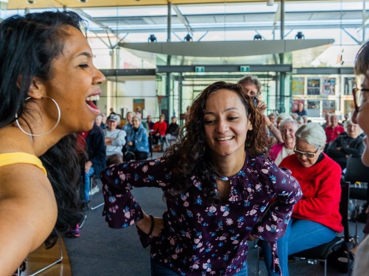 Image of Three women smile and laugh together in the foreground of a bright, modern room filled with seated people, suggesting a lively, joyful event or gathering.