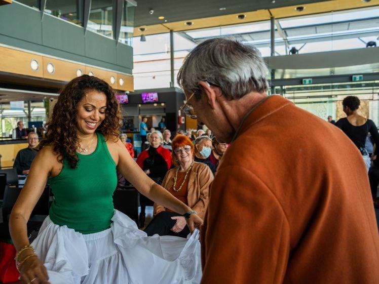 Image of A smiling woman in a green top and white skirt dances with an older man in an orange jacket indoors, whilst several people watch and smile in the background.