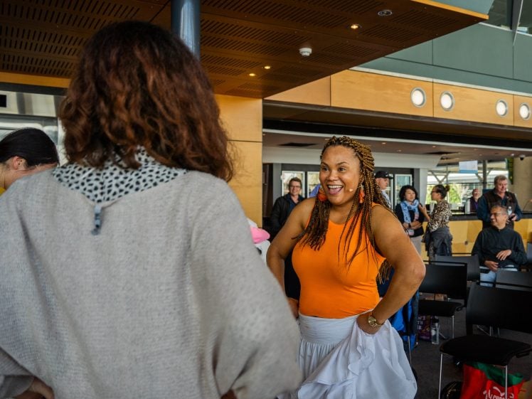 Image of Two women stand facing each other indoors, one smiling brightly in an orange top and white skirt, the other seen from behind. Several people are seated or standing in the background, watching the interaction.