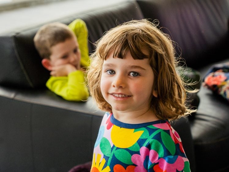 Image of A young child in a colourful flower-patterned shirt smiles at the camera, while another child in a green shirt reclines on a black sofa in the background.