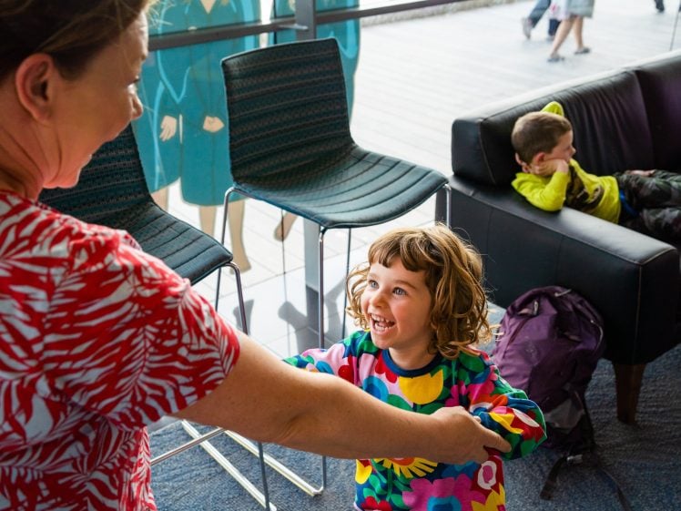 Image of A smiling child in a colourful shirt holds hands with an adult indoors, while another child in yellow relaxes on a sofa in the background near a purple rucksack.