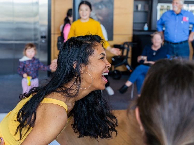 Image of A woman in a yellow top laughs joyfully, surrounded by people of different ages in a bright indoor space. Others in the background smile and watch her, creating a lively, cheerful atmosphere.