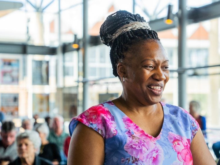 Image of A smiling woman in a floral top stands indoors, with a group of seated people and large windows in the background, giving the setting a bright, open atmosphere.