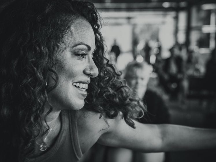 Image of A woman with curly hair smiles and stretches out her arm in a lively, indoor setting. The image is in black and white, with people blurred in the background.
