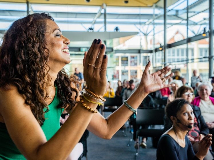Image of A woman in a green dress smiles and claps enthusiastically in a bright, modern room filled with seated people, some watching her and others looking in different directions.