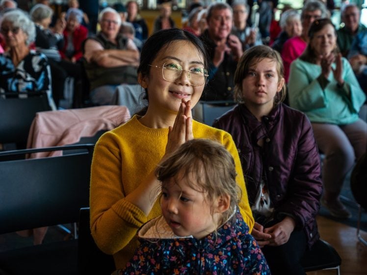 Image of A woman in a yellow jumper smiles and clasps her hands, sitting with a young girl on her lap among a diverse audience of people attentively watching an event in a bright indoor setting.