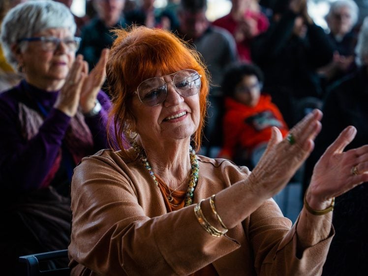 Image of An older woman with red hair, glasses, and a tan jacket is smiling and applauding at an event. Other people, seated and applauding, are visible in the blurred background.