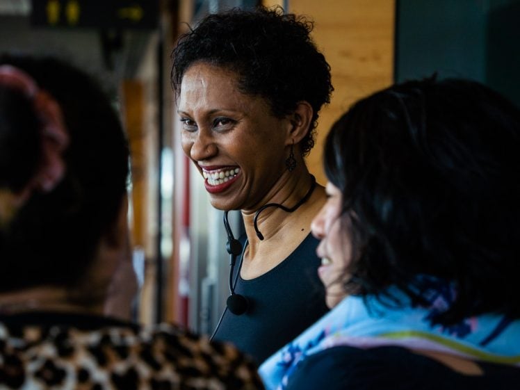 Image of A woman with short curly hair, wearing a black top and headset microphone, smiles and talks with two other women indoors. The atmosphere appears lively and friendly.