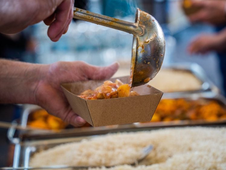 Image of A person uses a ladle to serve a steaming portion of stew or curry into a brown paper container, with trays of rice and other food dishes visible in the background.
