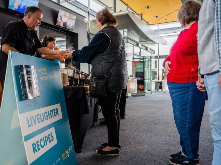 Image of People queue indoors at a food stall where a server hands a meal to a woman. A blue sign in the foreground reads LIVE LIGHTER RECIPES and FREE! Please take one. The setting appears to be a community event.