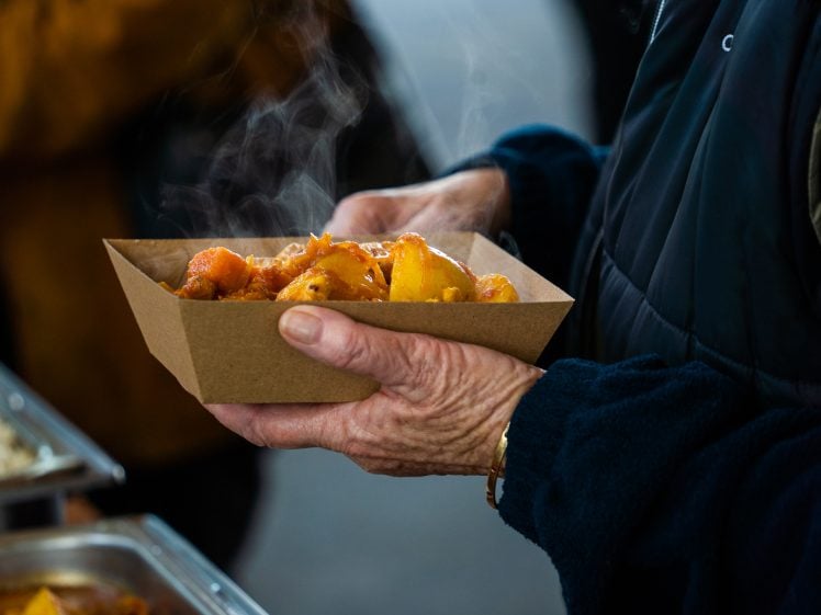 Image of A person holding a steaming serving of food in a brown paper tray, wearing a dark jacket and blue fleece, with trays of more food visible in the background.