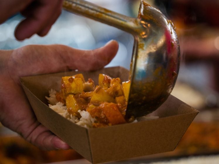 Image of A hand holds a cardboard tray filled with white rice as a metal ladle serves orange curry with chunks of vegetables or meat on top.