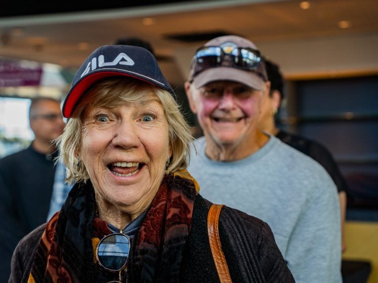 Image of An older woman in a baseball cap smiles excitedly at the camera, with a man in a cap and sunglasses on his head standing behind her, both indoors with blurred people and lights in the background.
