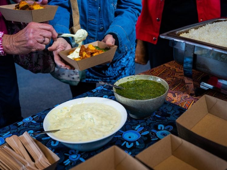 Image of People serving food onto cardboard trays at a buffet table with bowls of green sauce, creamy dip, a tray of rice, and disposable cutlery on a blue patterned tablecloth.