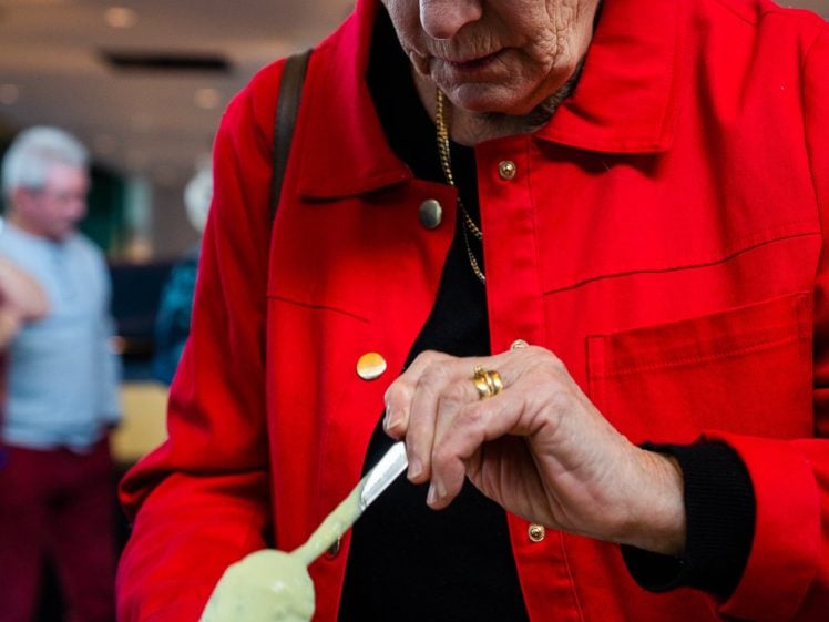 Image of An older woman in a red jacket and glasses spoons green sauce or cream onto food in a cardboard tray. She is focused on her meal, and there are people and lights blurred in the background.