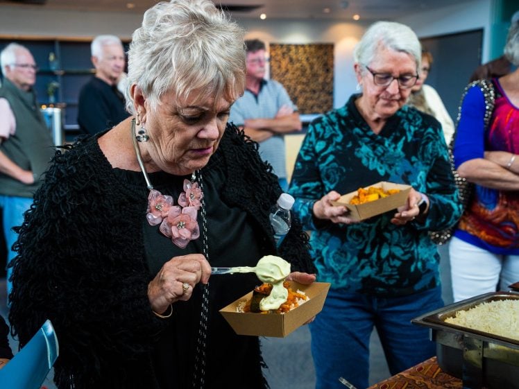 Image of An older woman tops her food with sauce at a buffet, whilst other older adults hold food trays and wait in queue in a brightly lit room.