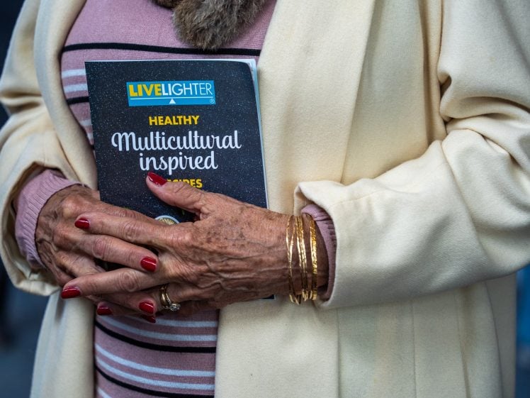 Image of An older woman with red painted nails and gold bangles holds a cookbook titled “Healthy Multicultural Inspired Recipes.” She wears a cream coat over a pink striped top.