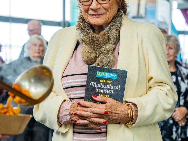 Image of An older woman in glasses and a cream coat holds a Healthy Multicultural Inspired Recipes booklet whilst standing by trays of food at a community event, with other people and large windows in the background.