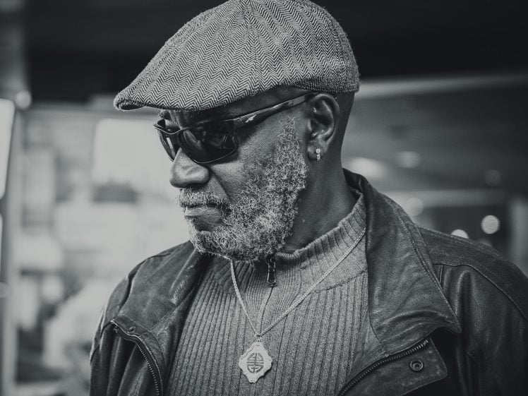 Image of A man with a grey beard wearing sunglasses, a flat cap, a leather jacket, and a necklace, looks to the side. The photo is in black and white with a blurred background.