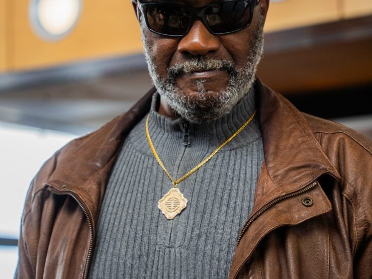 Image of A man wearing a grey flat cap, sunglasses, a gold necklace, a brown leather jacket, and a ribbed grey jumper, standing indoors with lights and wooden panels in the background.