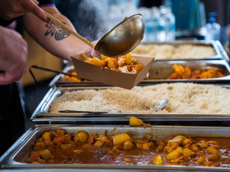 Image of A person ladles a serving of curry with potatoes into a cardboard tray above buffet trays filled with rice and curry, with steam rising from the hot food.