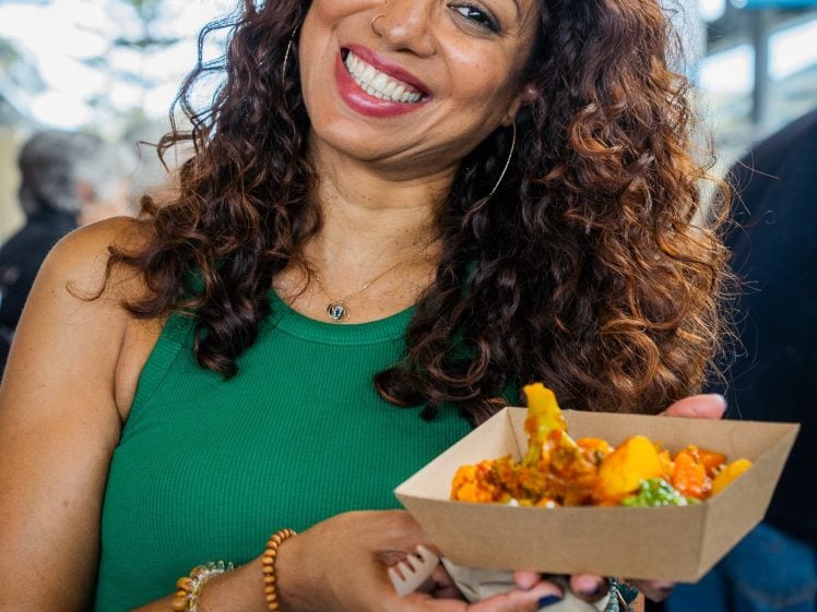 Image of A smiling woman with curly hair in a green sleeveless top holds a cardboard tray of colourful food with both hands at an indoor event.