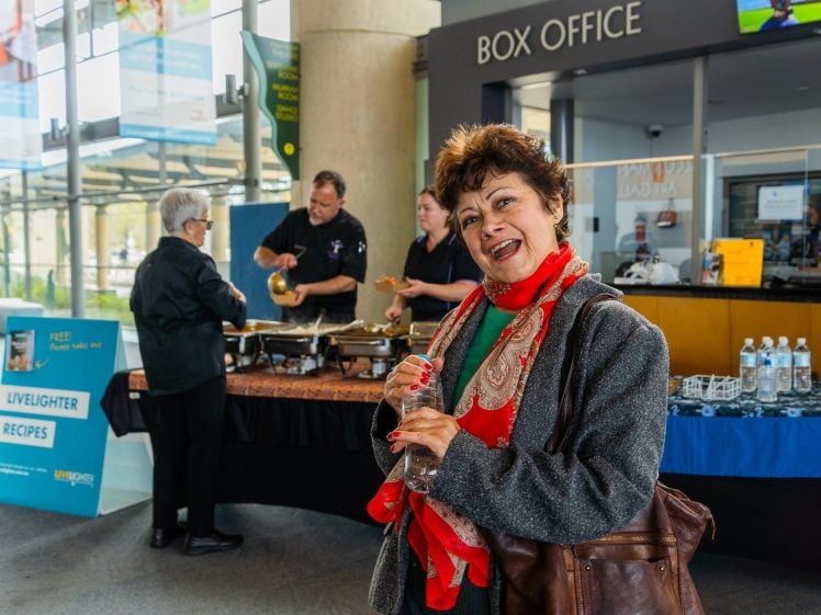 Image of A woman in a red scarf and grey coat smiles whilst holding a water bottle in front of a box office. Behind her, people are serving food at a table with chafing dishes and promotional signs.