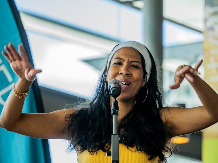 Image of A woman with long dark hair, wearing a white headband and yellow top, passionately sings or speaks into a microphone, gesturing expressively with both hands. Bright indoor setting with modern decor in the background.