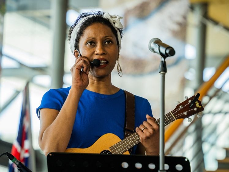Image of A woman wearing a blue shirt and white headband sings into a headset microphone whilst holding a ukulele, standing in front of another microphone with a blurred indoor background.