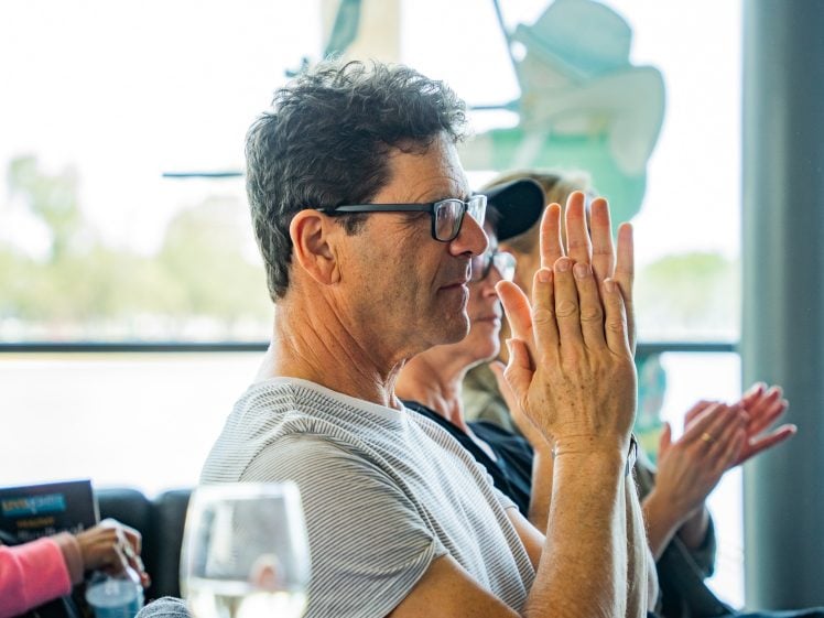 Image of A man wearing glasses and a striped shirt sits indoors, facing left and clapping. Other people in the background are also applauding. There is a window behind them letting in natural light.