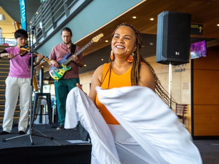 Image of A smiling woman in an orange top and white skirt dances in the foreground whilst two musicians play instruments on a stage behind her in a bright, modern indoor setting.