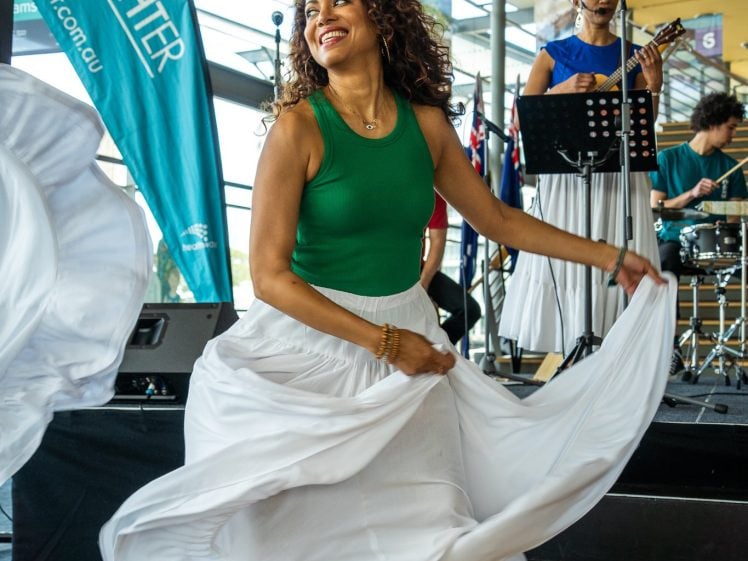 Image of A barefoot woman in a green top and flowing white skirt dances joyfully indoors, holding her skirt as musicians perform on stage behind her. A teal LiveLighter banner stands to the left.