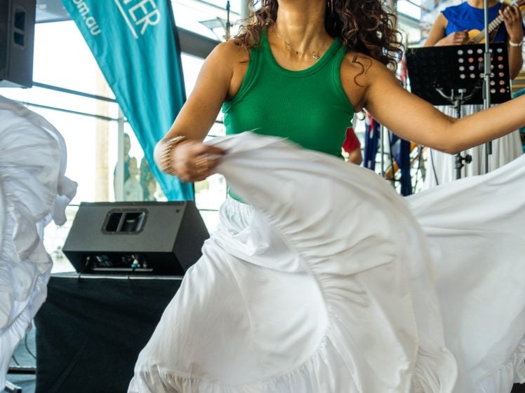 Image of A woman in a green top and flowing white skirt joyfully dances indoors, smiling with arms outstretched. Musicians play in the background. A LiveLighter banner and speakers are visible behind her.