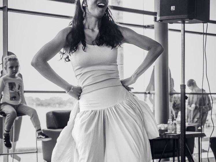 Image of A woman in a flowing white skirt and vest top dances joyfully indoors, smiling with her hands on her waist. In the background, a child sits on a ledge and large windows let in natural light.