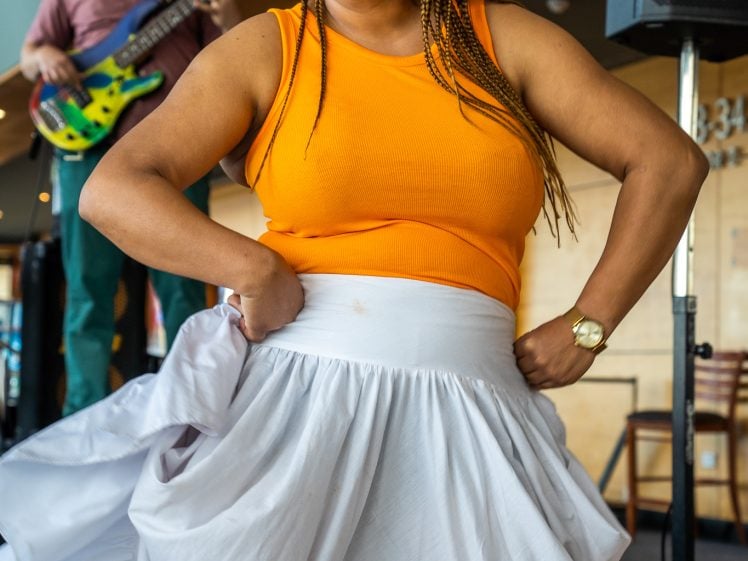 Image of A woman in an orange sleeveless top and long white skirt dances onstage, smiling with her hands on her hips, whilst a musician plays guitar in the background.