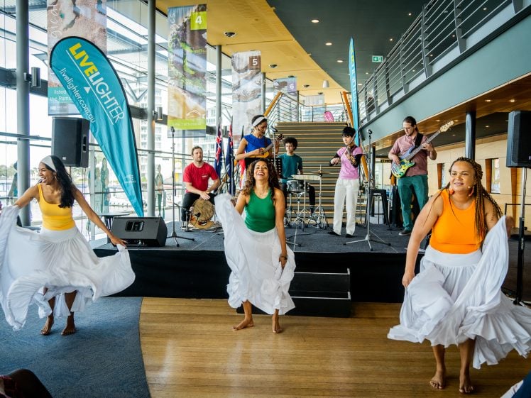Image of Three women in colourful tops and white skirts dance energetically in front of a live band performing on a stage in a modern, bright indoor venue with large windows and banners.