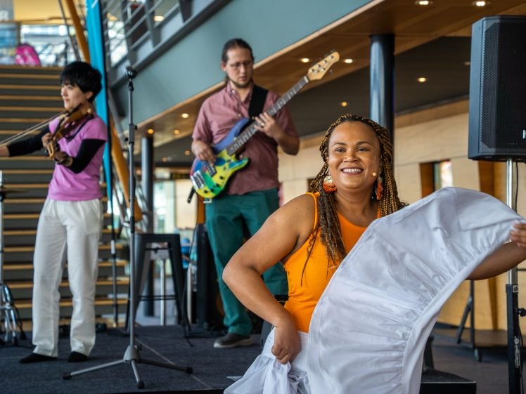 Image of A smiling woman in an orange top dances with a white skirt on stage, while three musicians play violin, bass guitar, and flute in the background. The setting appears to be indoors with stairs and railings behind them.