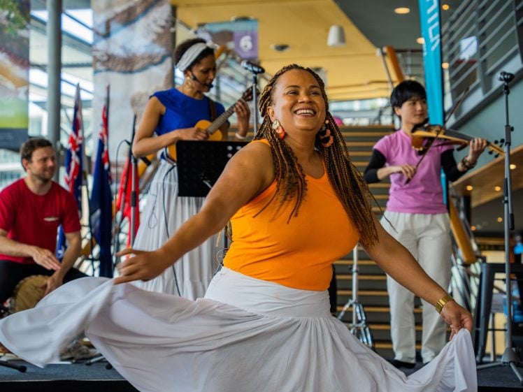Image of A smiling woman in an orange top and white skirt dances in front of a band. Three musicians play a drum, ukulele, and violin on a stage with flags and microphones in the background.