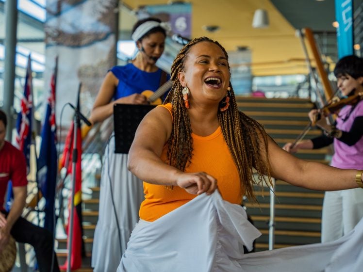 Image of A joyful woman in an orange top dances whilst three musicians play instruments in the background. Flags are displayed to the side, and the scene is set indoors with stairs and bright natural light.
