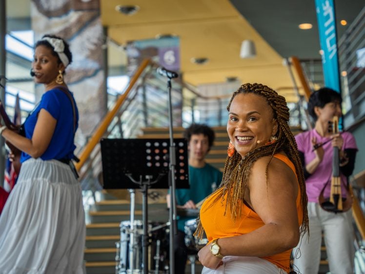 Image of A smiling woman in an orange top stands in the foreground whilst musicians play instruments behind her on an indoor stage with stairs and modern decor in the background.