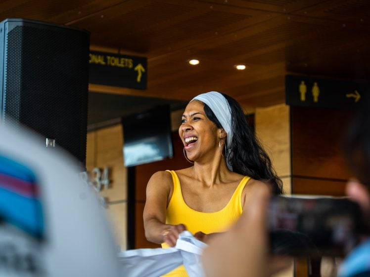 Image of A woman in a yellow top and white headband smiles brightly whilst dancing indoors, with people in the foreground and toilet signs visible in the background.