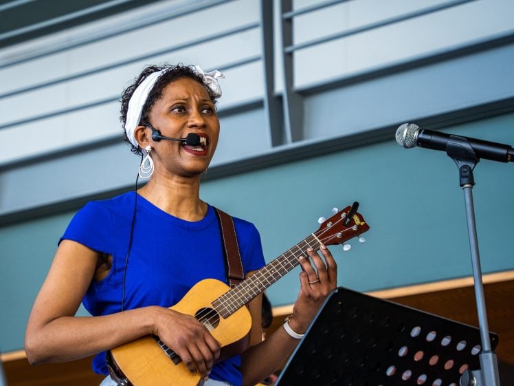 Image of A woman wearing a blue shirt and a white headband sings into a headset microphone while playing a ukulele. She stands near a microphone stand and music stand, performing indoors.